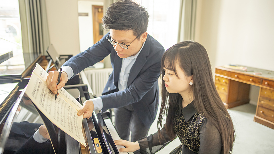 An Asian man teaching a female Asian student, practising on the piano.